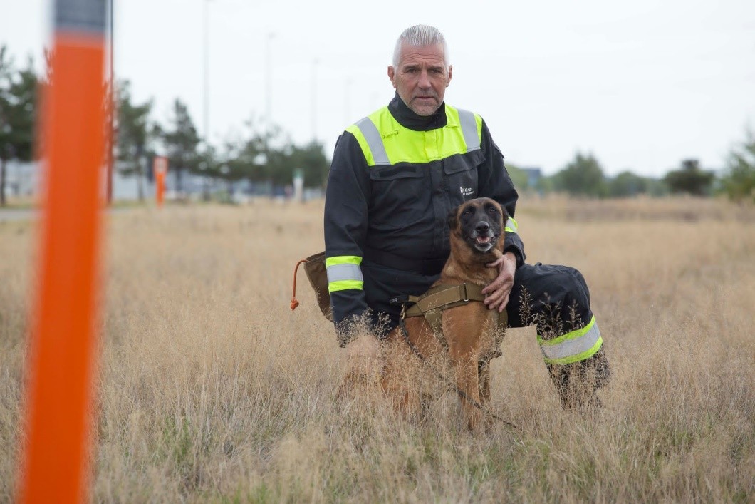 K9Belgium.com handler in high-visibility gear with a Malinois at a marker post on an industrial site